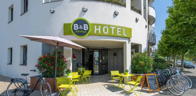 La facade extérieure de l"hôtel, avec des tables et chaises métalliques vertes devant.